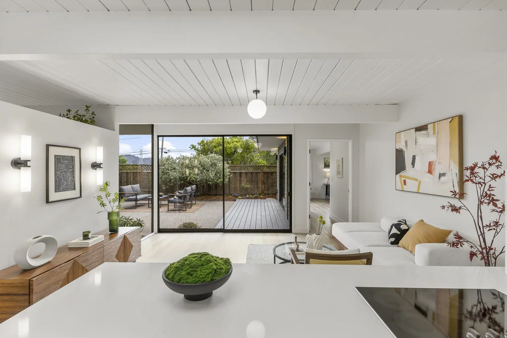 Looking across a kitchen counter through the living room to a sliding glass wall opening onto a rear deck and fenced garden.